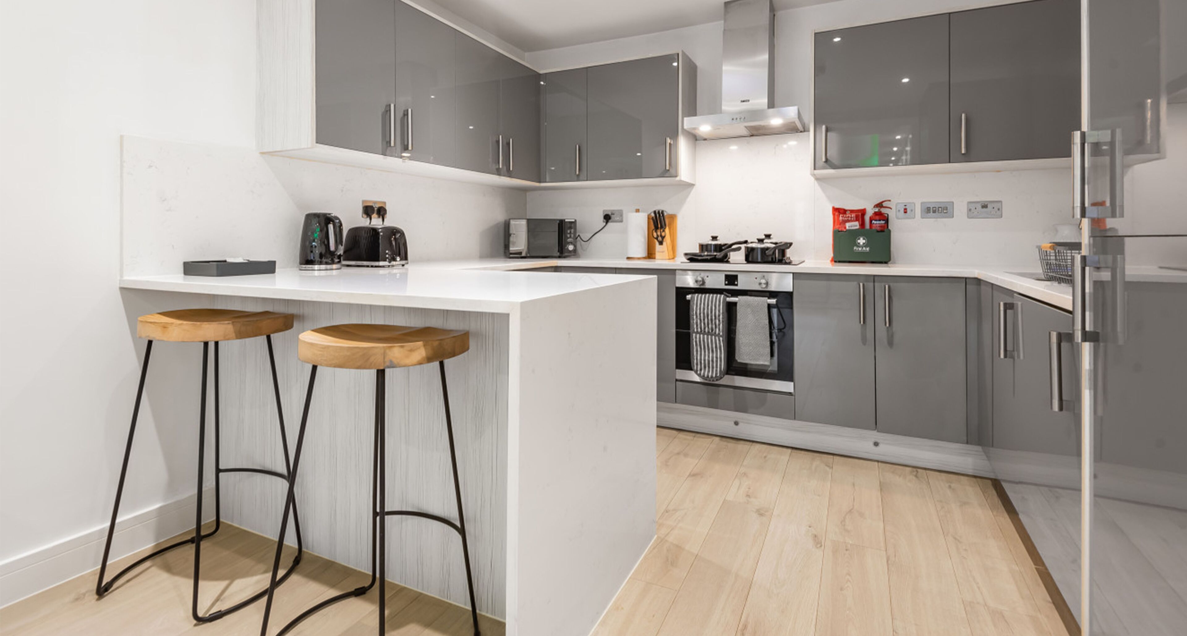 A modern kitchen with grey cabinets, a white countertop, two wooden stools, a built-in oven, and a refrigerator.