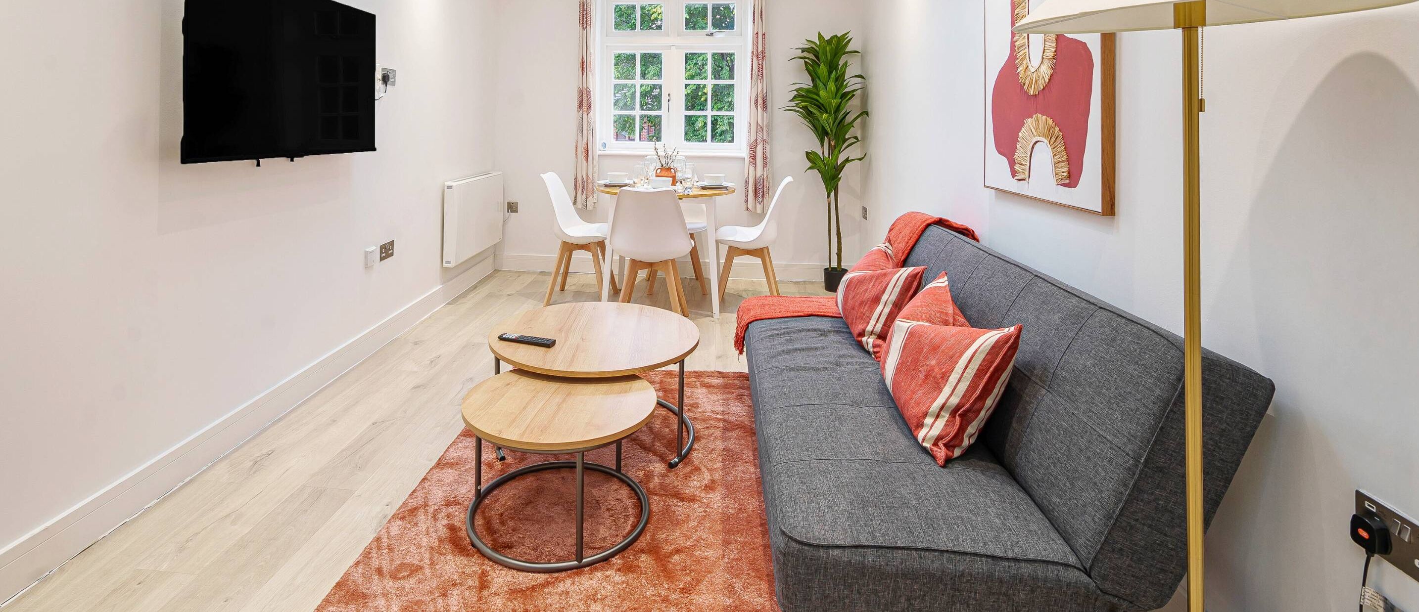 A modern living room with a grey sofa, two round wooden tables, a TV mounted on the wall, and a dining area with a white table and chairs.