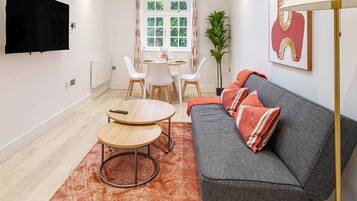 A modern living room with a grey sofa, two round wooden tables, a TV mounted on the wall, and a dining area with a white table and chairs.