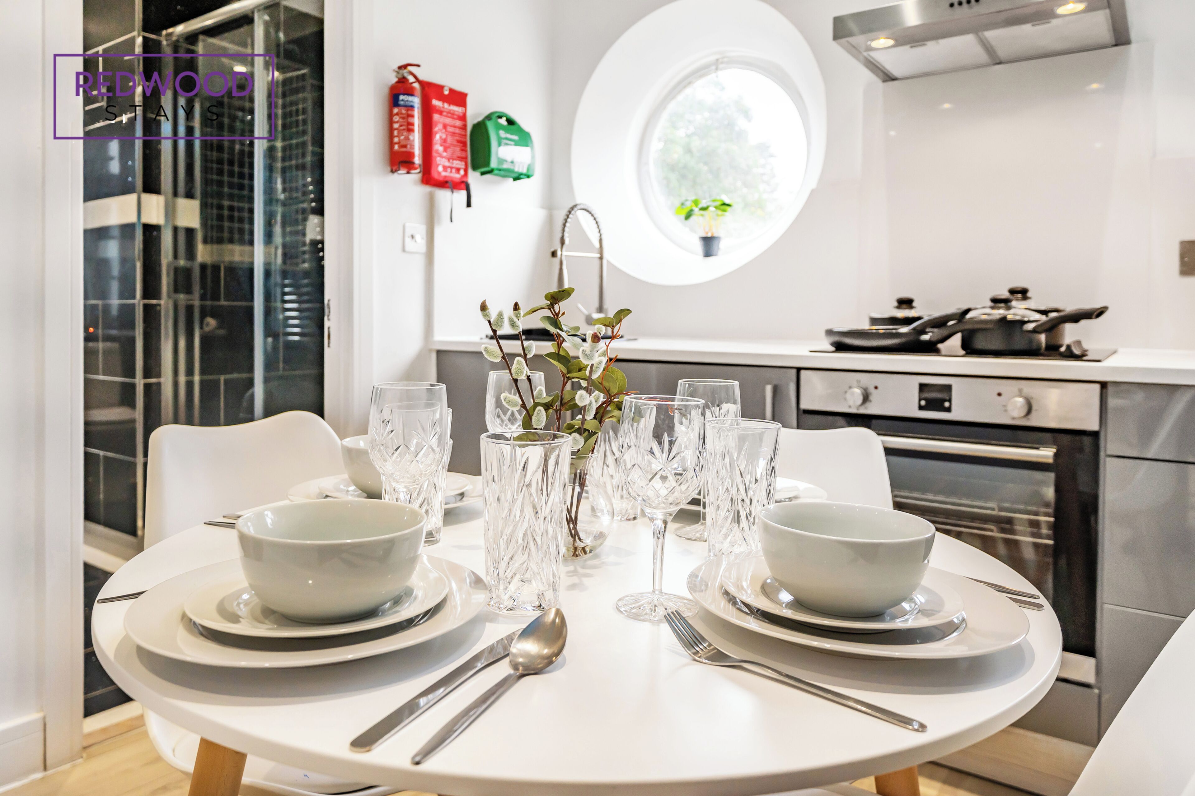 Una cucina moderna con un tavolo da pranzo apparecchiato per due, dotata di ciotole, piatti e posate bianche. Sul tavolo c'è un vaso con fiori e una finestra lascia entrare luce naturale.