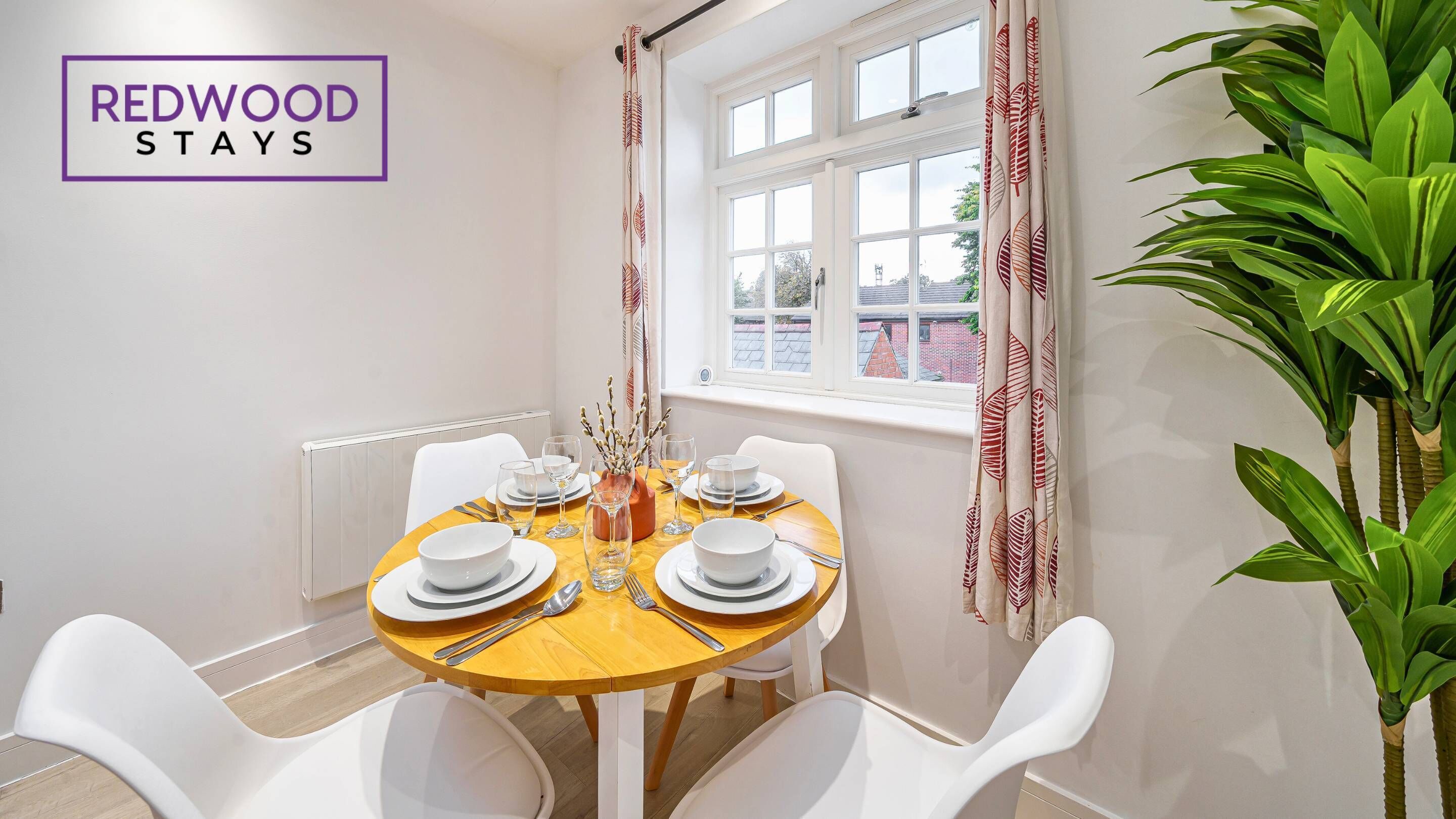 A dining area with a round table set for four, white chairs, a window with curtains, and a plant on the right.