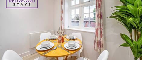 A dining area with a round table set for four, white chairs, a window with curtains, and a plant on the right.