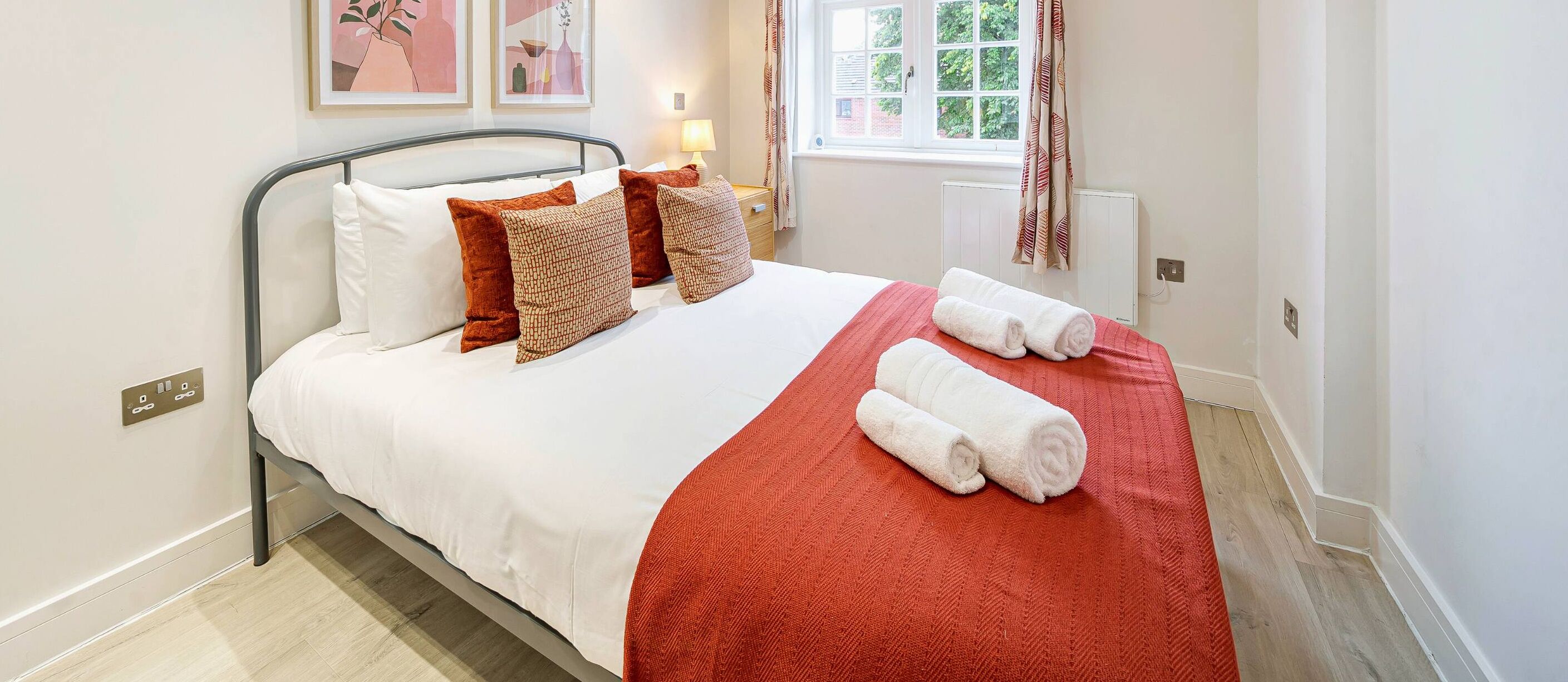 A neatly made bed with white and red bedding, a red throw blanket, and rolled towels.