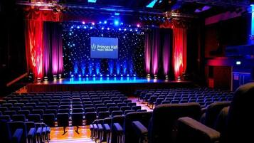 An auditorium with rows of seats, a stage with a large screen displaying "Pringles Hall Theatre - Aldenhot," and red curtains.