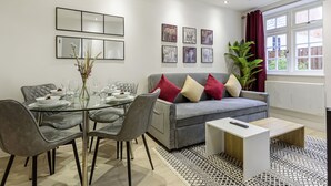 A modern living room with a dining area, grey furniture, a grey sofa with red and beige cushions, a small white coffee table, and a large window with curtains. - Ragan Court Apartments in Aldershot (Aldershot)
