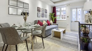 A modern dining area with a round glass table, grey upholstered chairs, and a grey sofa with red and white cushions. - Ragan Court Apartments in Aldershot (Aldershot)