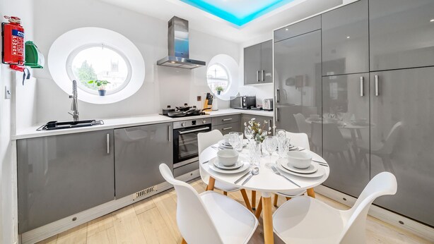 A modern kitchen with a round dining table set for four, white chairs, a built-in oven, and a window with a view of buildings. - Ragan Court Apartments in Aldershot (Aldershot)