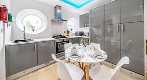 A modern kitchen with a round dining table set for four, white chairs, a built-in oven, and a window with a view of buildings. - Ragan Court Apartments in Aldershot (Aldershot)
