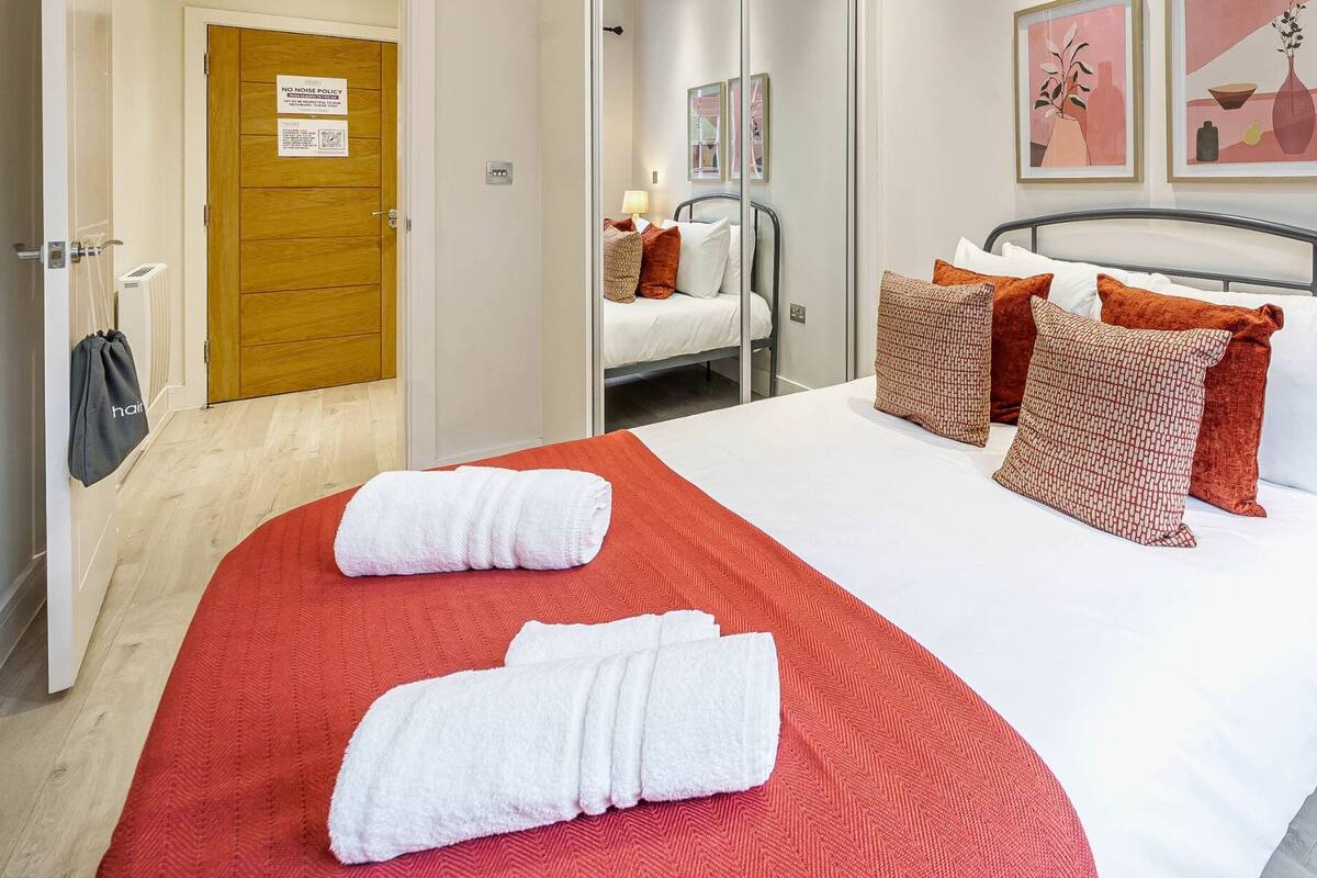 A neatly made bed with white and red linens, a wooden nightstand with a lamp, and a door with a 'No Noise Policy' sign.