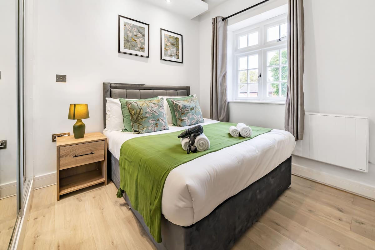 A bedroom with a bed, a nightstand, a lamp, a window with curtains, and two framed pictures on the wall.