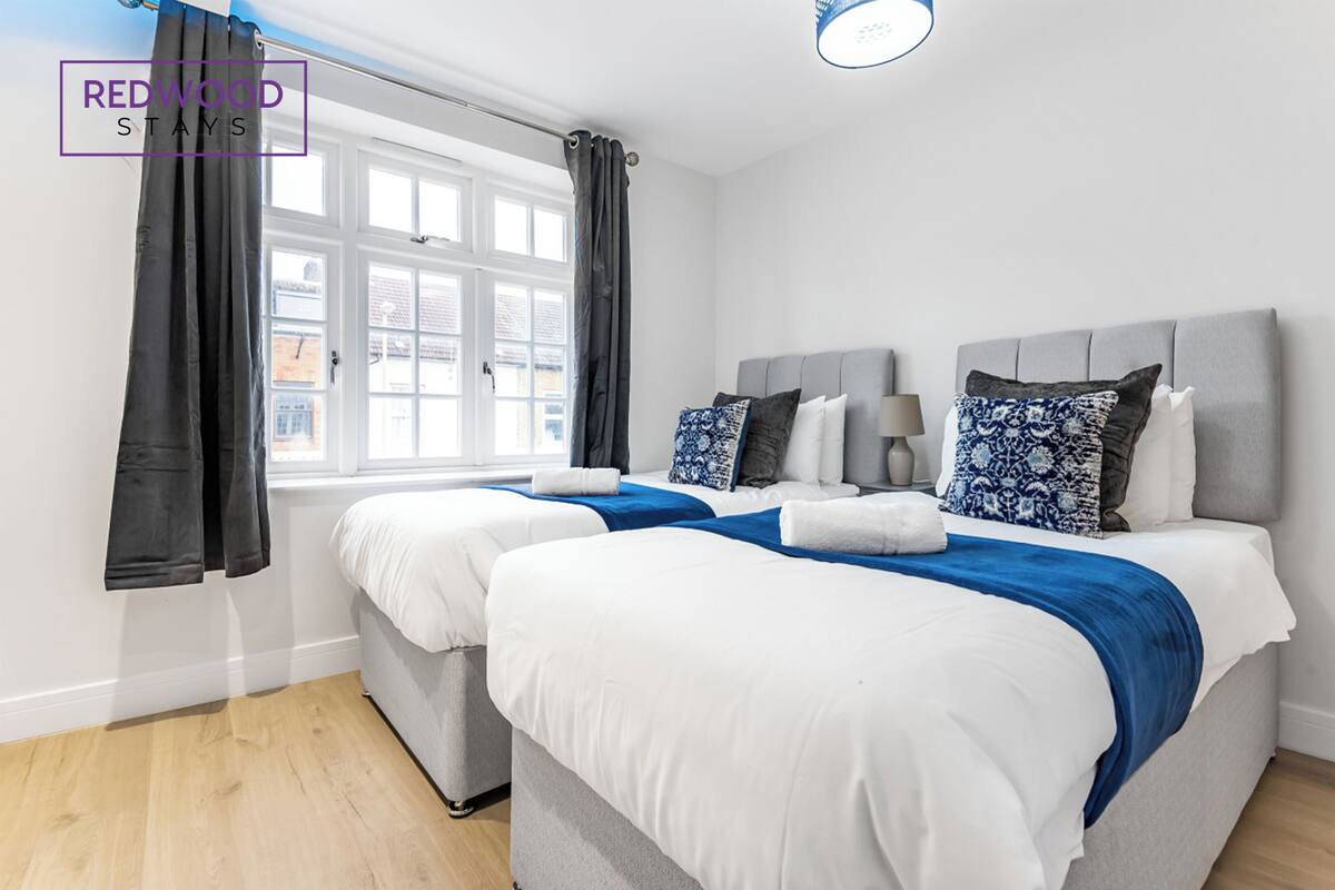 A hotel room with two beds, a window with curtains, a lamp, and a view of buildings outside.