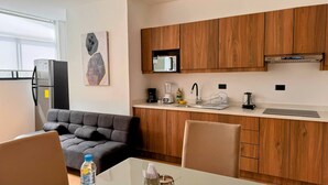 Una cocina compacta con armarios de madera, microondas, cafetera y licuadora. Hay un sofá gris y una mesa de comedor con sillas.