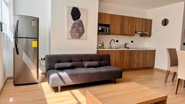 Un espacio habitable pequeño con kitchenette, un sofá gris, una mesa de centro de madera y un reloj moderno en la pared.