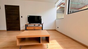 Una habitación minimalista con una mesa de centro de madera, un televisor de pantalla plana sobre un mueble, una puerta de madera oscura y una ventana con vista a un edificio.