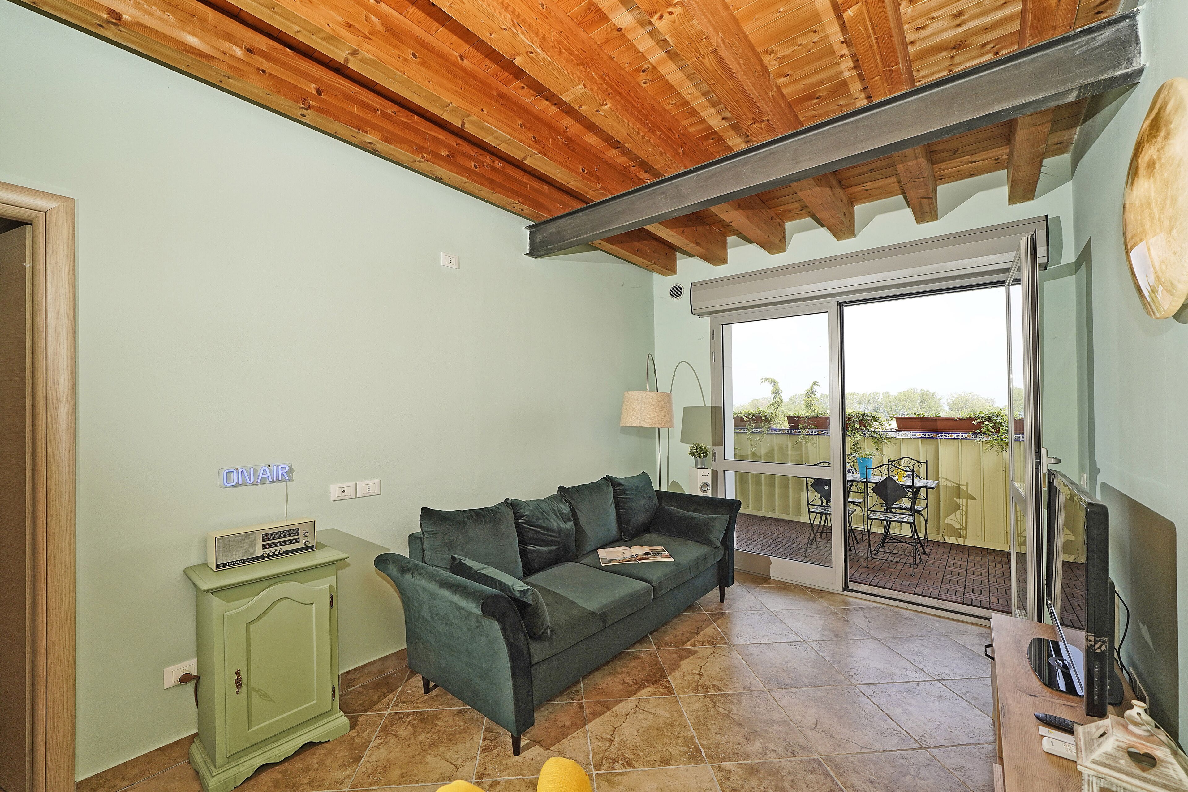 Un salon avec un canapé vert, un plafond en bois, un téléviseur et une porte-fenêtre coulissante donnant sur une terrasse extérieure.