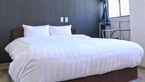 Una cama bien hecha con sábanas blancas en una habitación de hotel con pared gris y una mesita con una silla.