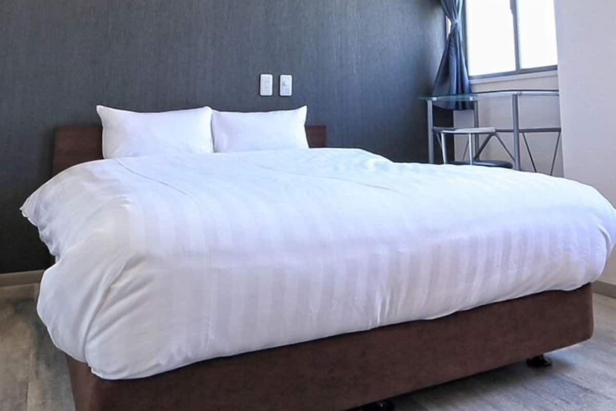 Una cama bien hecha con sábanas blancas en una habitación de hotel con pared gris y una mesita con una silla.