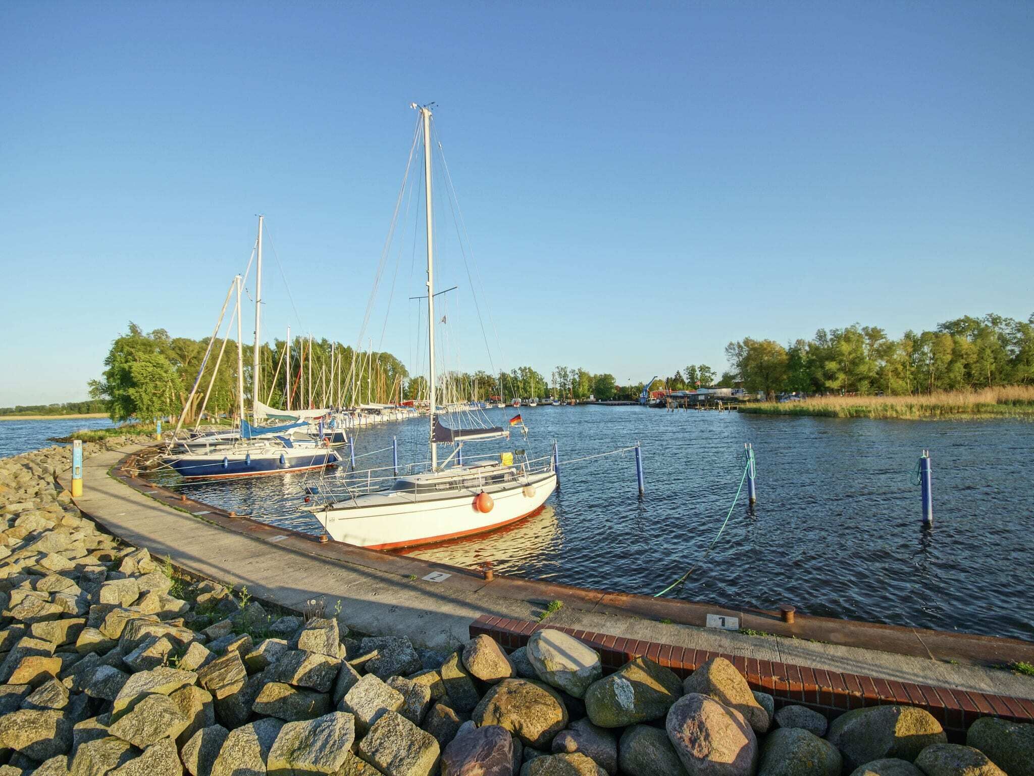 A marina with several sailboats docked, a concrete walkway, and a rocky shoreline.