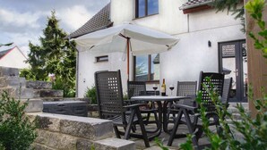 Een terras buiten met een tafel, stoelen, een witte parasol en een fles wijn.