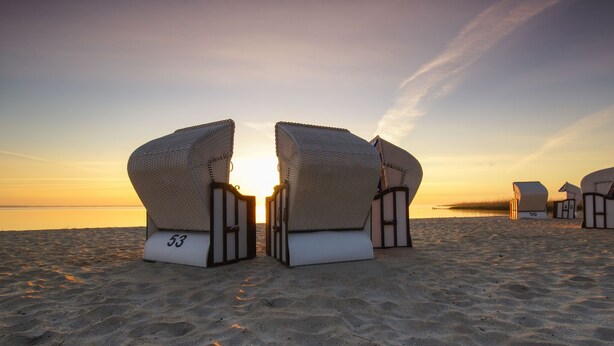 Strandstoelen met nummers 53 en 54, en een gebouw met nummer 50 op een zandstrand bij zonsondergang.