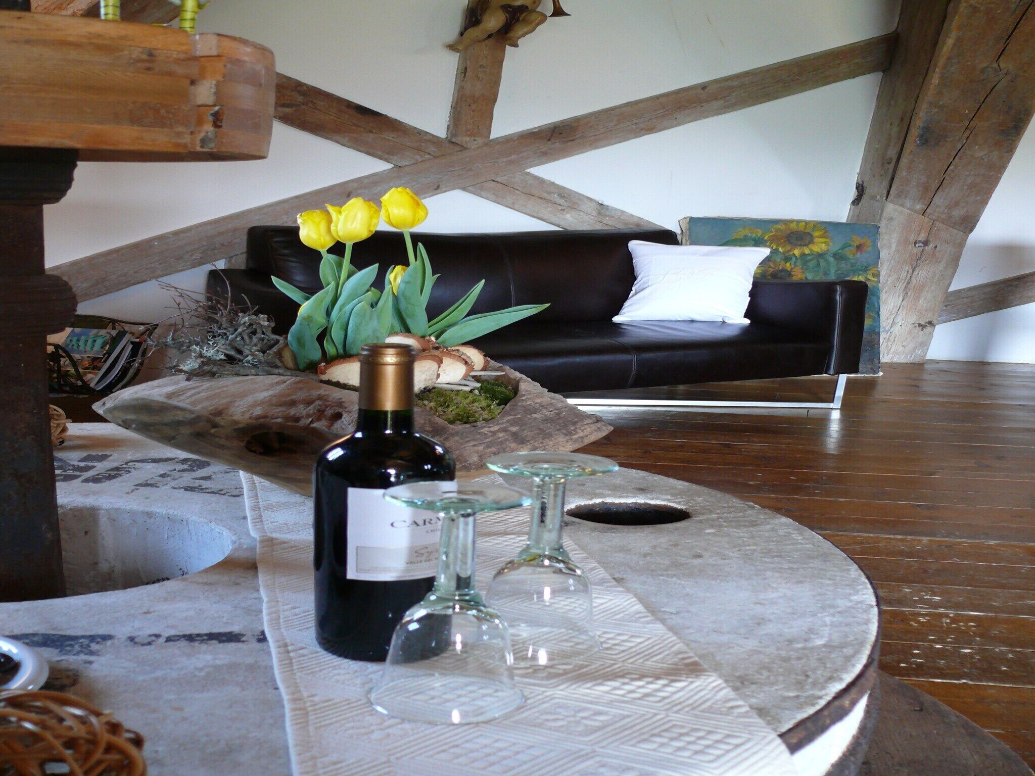 Un accogliente soggiorno con soffitto in legno, un divano nero con un cuscino bianco, un tavolino in legno con una bottiglia di vino e due bicchieri, e un vaso con fiori gialli.