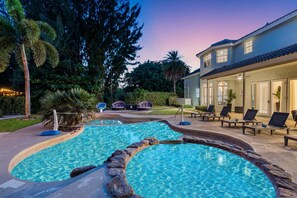 A pool with a natural rock border, lounge chairs, and a building with large windows and a covered patio area. - 8BR Entertainers Estate w/ Pool, Games & Theater ! (Coconut Creek)