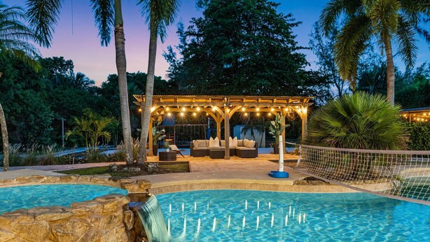 A pool with a waterfall feature, a covered seating area with lights, and surrounding tropical vegetation. - 8BR Entertainers Estate w/ Pool, Games & Theater ! (Coconut Creek)