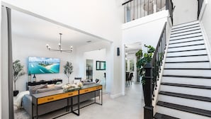 A modern living room with a large TV, a sofa, a coffee table, and a staircase. - 8BR Entertainers Estate w/ Pool, Games & Theater ! (Coconut Creek)