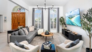 A modern living room with a grey sofa, a coffee table, and a TV displaying a cityscape. - 8BR Entertainers Estate w/ Pool, Games & Theater ! (Coconut Creek)