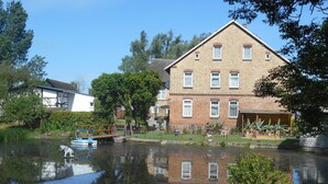 A riverside building with a brick facade, a dock with a small boat, and a reflection in the water. - Vacation Home with Pond (Klein Pravtshagen)