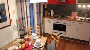 Eine Küche mit weißen Schränken, einem Herd und einer Mikrowelle. Es gibt einen Esstisch mit einer roten Tischmatte, Äpfeln und einer Schüssel mit Essen.