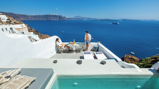 A rooftop pool with a clear view of the sea, white buildings, and a person standing by the pool.