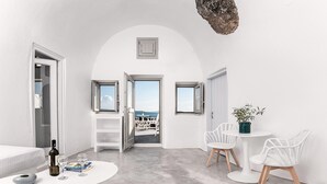 A modern, minimalist room with white walls and a concrete floor. It features a dining area with a round table and white chairs, a small white sofa, and a window with a view of the sea. - Suite with Private Hot Tub (Santorini)