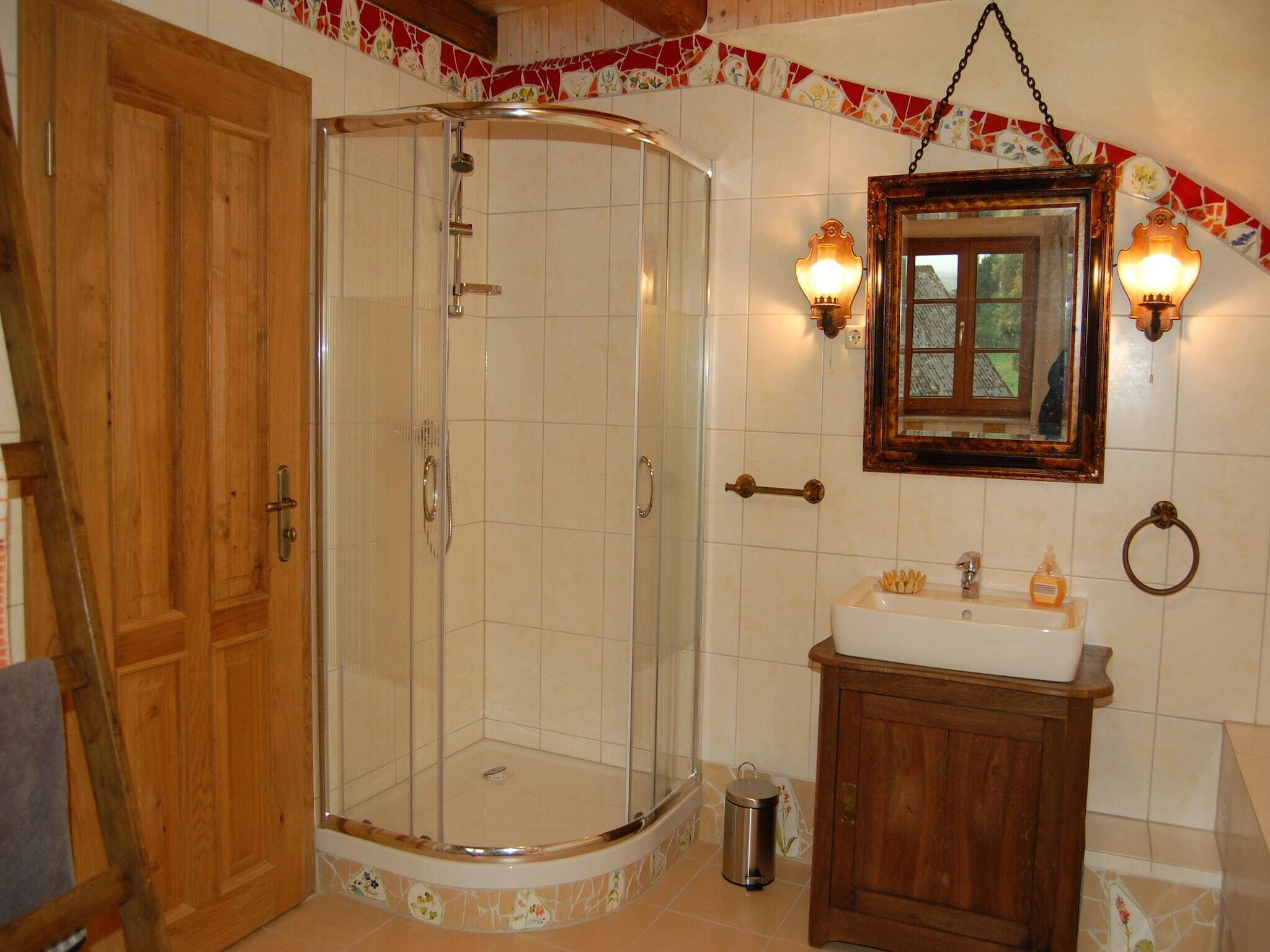 A bathroom with a glass shower enclosure, wooden door, tiled walls, a wooden cabinet, and a mirror with wall-mounted lights.