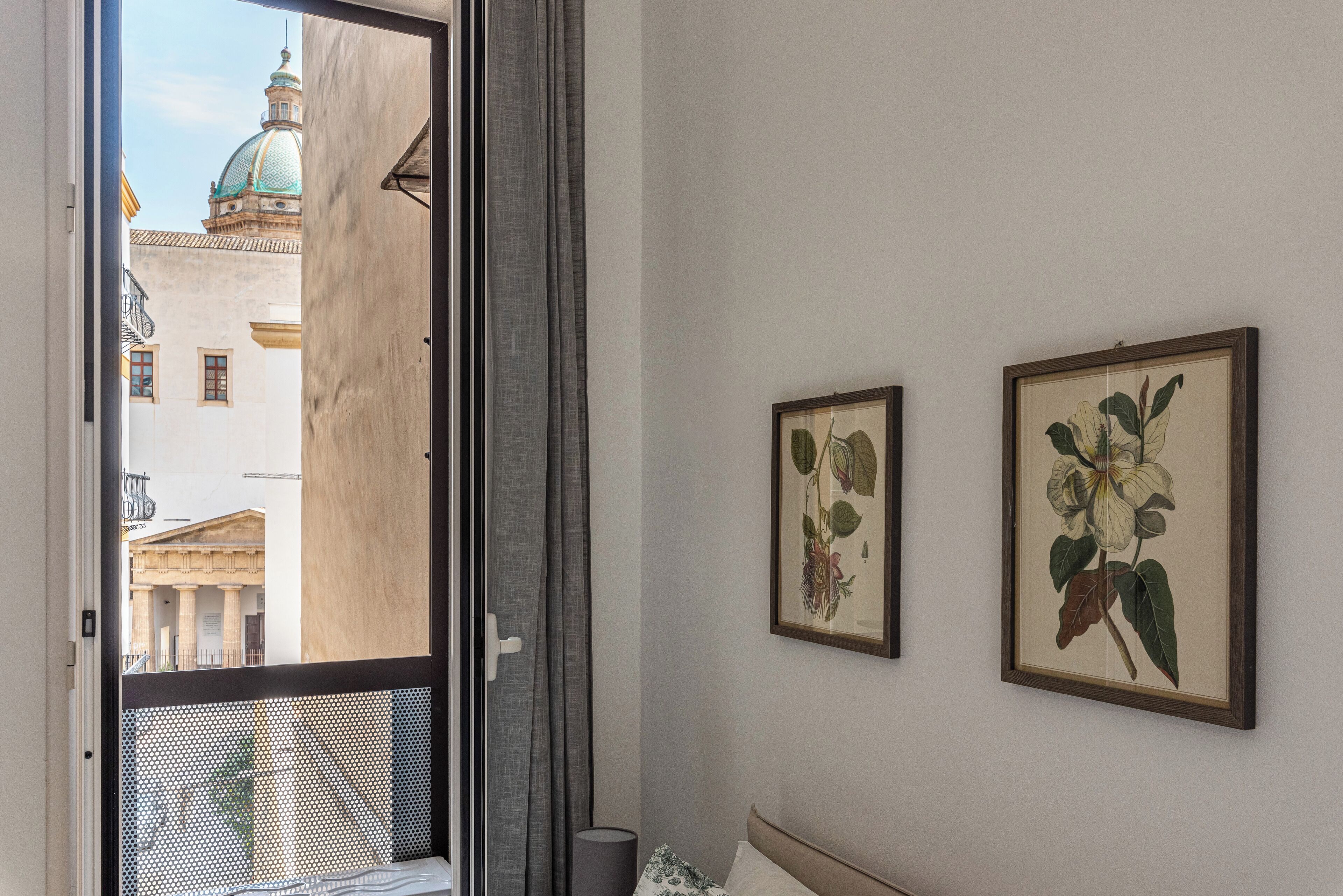A room with a bed, a window with a view of a building, and two framed botanical prints on the wall.