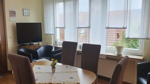 Un'area pranzo con un tavolo rotondo, sei sedie, una televisione e una finestra vista su un tetto in tegole rosse e vegetazione.