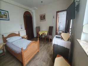 Une chambre à coucher avec un lit en bois, une petite table recouverte d’une nappe jaune, une chaise et un bureau avec une lampe.