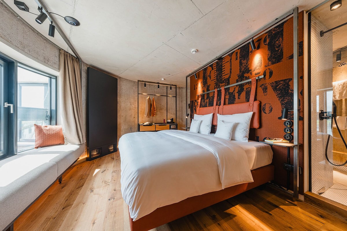 Una habitación de hotel moderna con una cama grande, un ventanal con vistas, suelo de madera y un diseño de pared único.