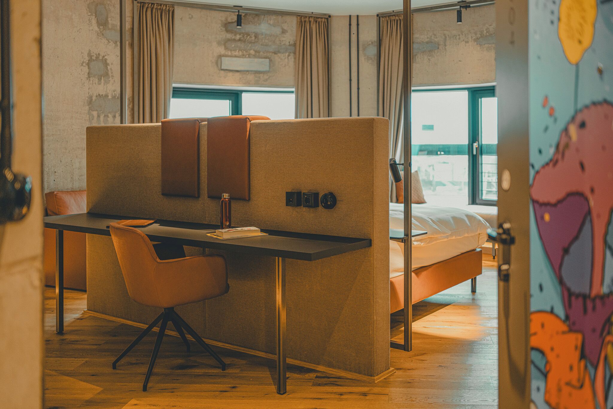 A compact room with a bed, a desk, and a chair. There is a window with curtains, a wall-mounted light, and a door with a colorful mural.