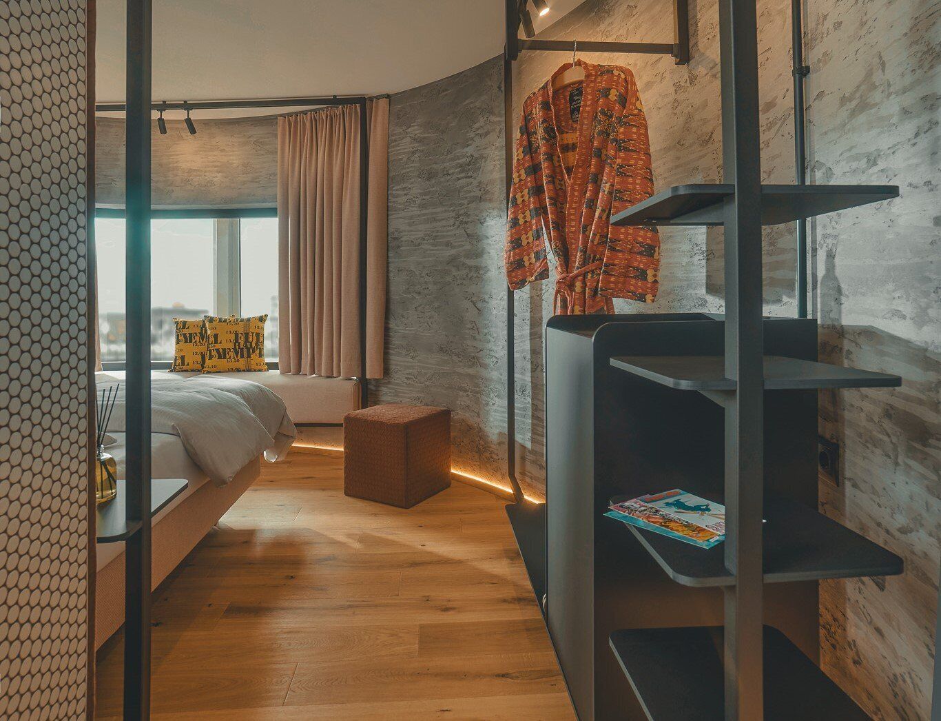 A modern bedroom with a bed, a small stool, a shelf with books, and a window with curtains.