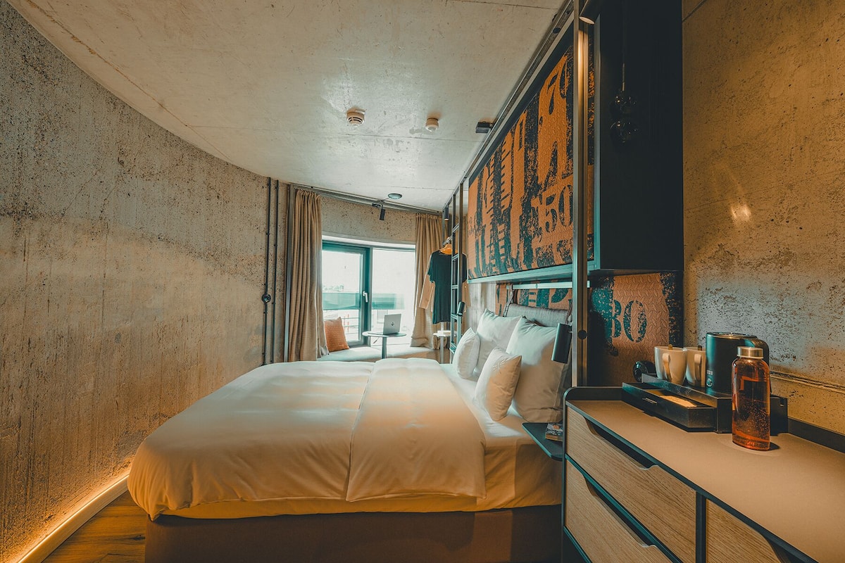 Habitación de hotel con una cama grande, un escritorio con cafetera y vistas a la ciudad.