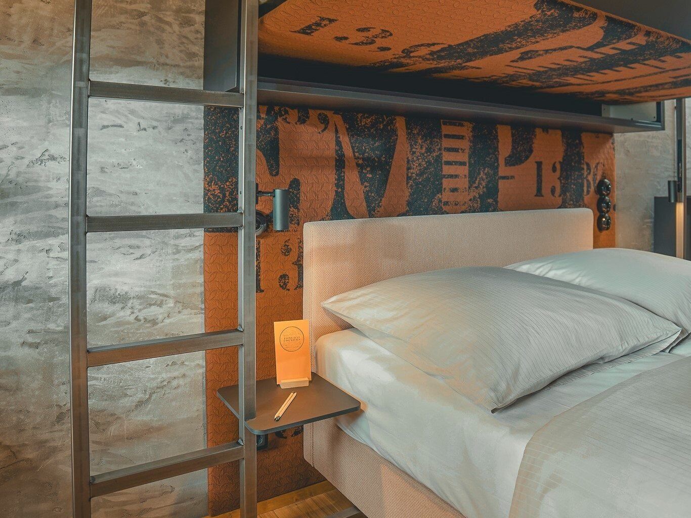 Uma cama com cabeceira encostada em uma parede texturizada, uma escada e uma pequena mesa com um livro.