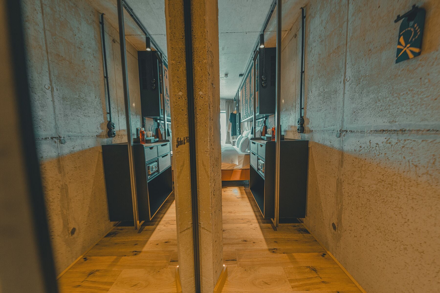 A narrow corridor with wooden flooring leading to a room with a bed, a desk, and a chair.