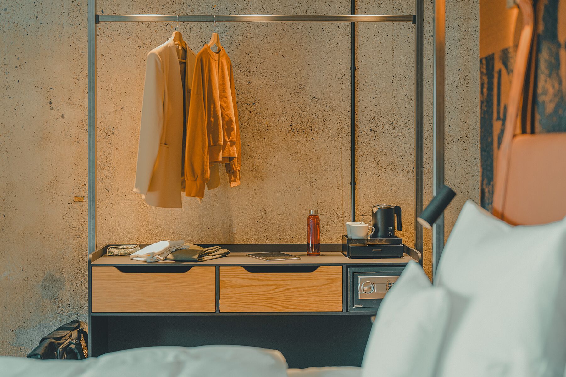 A hotel room with a bed, a bedside table with a coffee maker, a mirror, and clothes hanging on a rack.