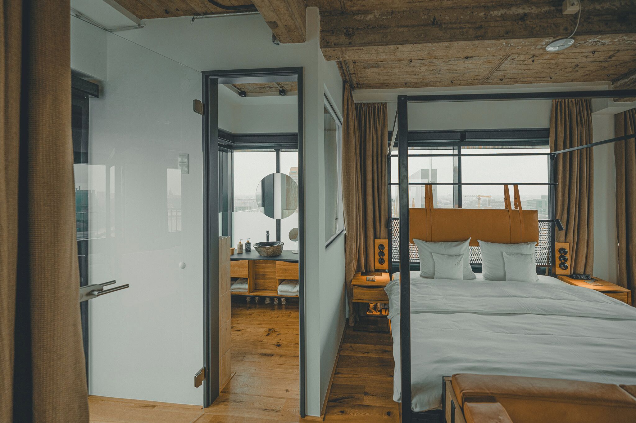 A modern hotel room with a wooden ceiling, a large bed, a desk with a mirror, and a view of the city through large windows.