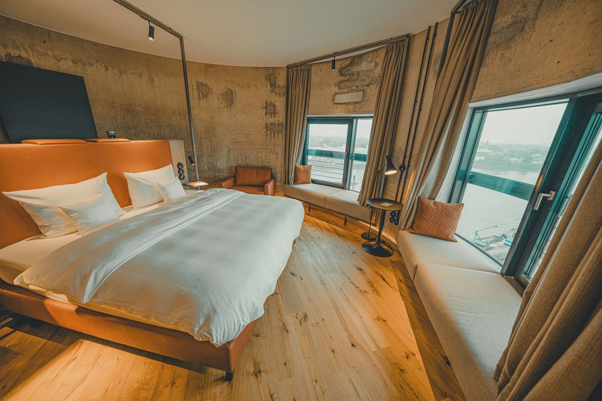 Habitación de hotel moderna con una cama grande, un sofá, una mesita y vistas al agua a través de amplios ventanales.