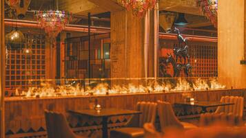 A restaurant interior with wooden tables and chairs, a decorative chandelier, and a fire feature along the back wall.