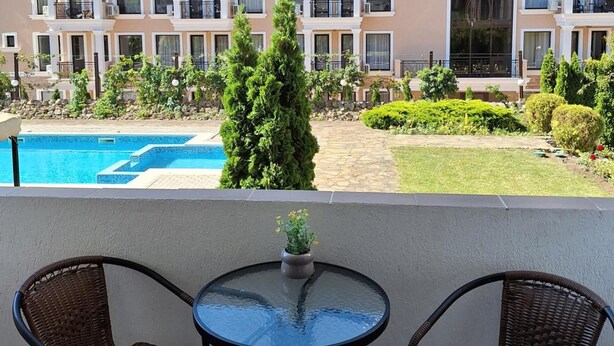 Un balcon avec deux fauteuils en osier et une petite table, donnant sur une piscine et un jardin bien entretenu.