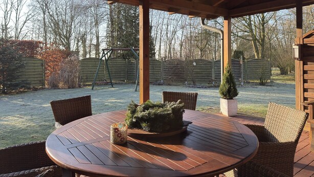 Un patio techado con una mesa redonda de madera, sillas de mimbre y una pequeña planta en maceta.