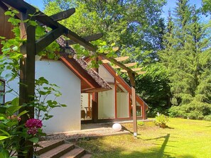 Una casa con una pérgola de madera, un jardín con césped y una planta en maceta, y un pequeño camino de piedra que lleva a la entrada.